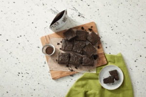 Black cocoa brownies sit on top of a wooden cutting board. Nearby is a plate with a half eaten brownie on top of a green towel and an open bag of black cocoa.