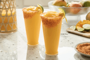 Two chili-mango margaritas with spice on their rims stand on a counter surrounded by plates and bowls holding fruit.