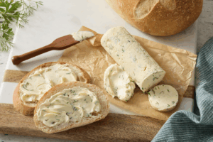 Homemade bread slices with large amounts of fancy butter. Nearby is a log of fancy butter, a loaf of bread and a wooden butterknife. All items sit on a cutting board.
