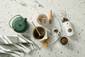 A bowl of Hojicha surrounded by objects such as an ornate tea pot, sieve and towel. All items sit on a black and white counter.