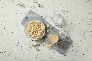 A bowl of macadamia nuts near a container of macadamia butter, a cup of milk and a butterknife. All items sit on a long, grey cutting board.