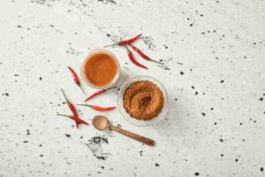A small bowl of piri piri sauce next to a larger bowl of piri piri powder, multiple piri piri peppers and a wooden spoon.