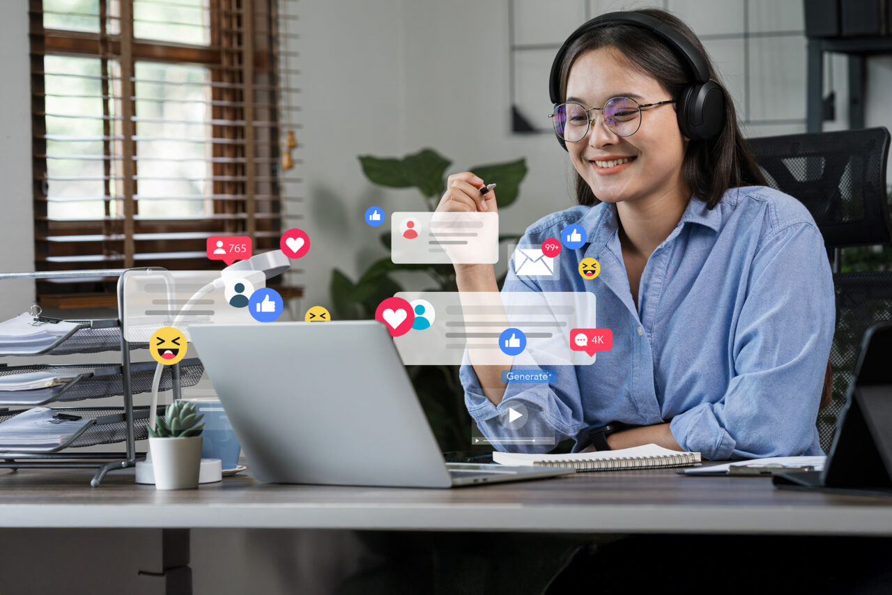 A woman wearing glasses and headphones sits at a table using a laptop. Floating between her and the laptop are icons showing social media posts and post reactions such as “like” or “heart” emojis.